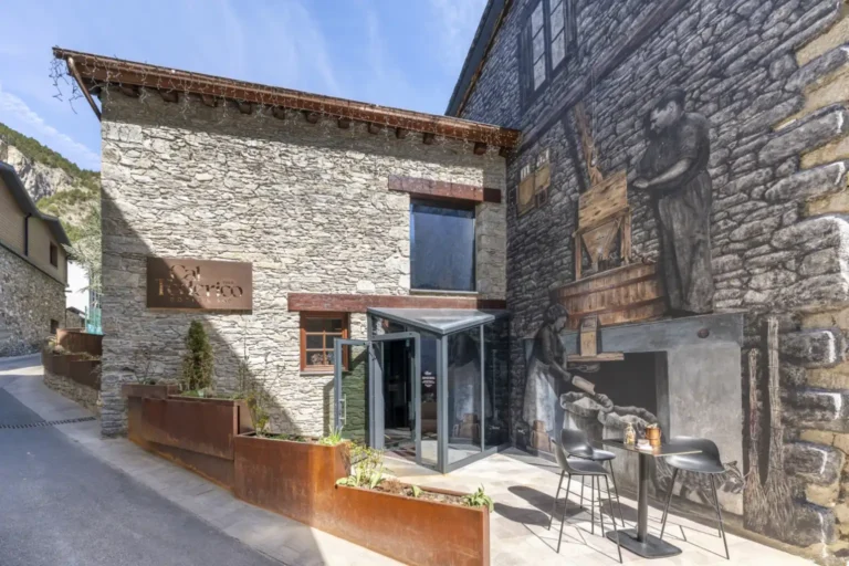Façana principal de Cal Federico amb el rètol, el vestíbul vidriat d'accés i el mural mural que decora el mur lateral de pedra.